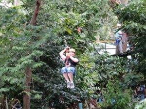 Lynn approaching a zipline platform through the rainforest canopy with a guide watching from the platform behind her.