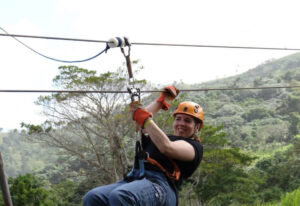 Lynn smiling while ziplining above the rainforest in Punta Cana, wearing an orange helmet and harness with green hills behind her.