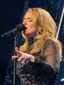 A singer performing live on stage in a sparkly black dress, reaching out to the audience under blue stage lighting.