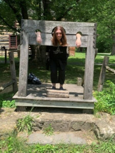 A person with long hair is smiling and standing in wooden stocks, an old punishment device, outdoors—a scene that sparks curiosity—in a grassy, sunny area with trees and a rustic wooden building in the background.