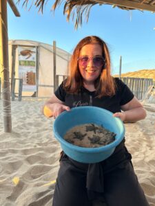 Lynn kneeling in the sand holding a blue bowl filled with tiny sea turtle hatchlings in wet sand, with the greenhouse and palm roof shelter behind her.