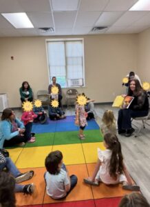 Lynn A. Haller reads The Hallway of Doorknobs to a group of children seated on a rainbow carpet at the Tyrone-Snyder Public Library. Children's faces are covered with sun stickers.